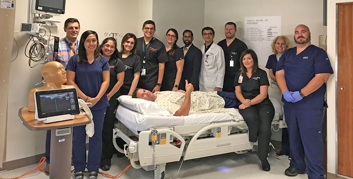 The Neurocritical Care Team standing around a hospital bed smiling while they practice invasive line placement.