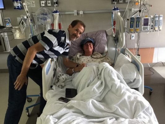 Carolyn Taylor sitting in a hospital bed and smiling. She is surrounded by machines and hospital equipment.