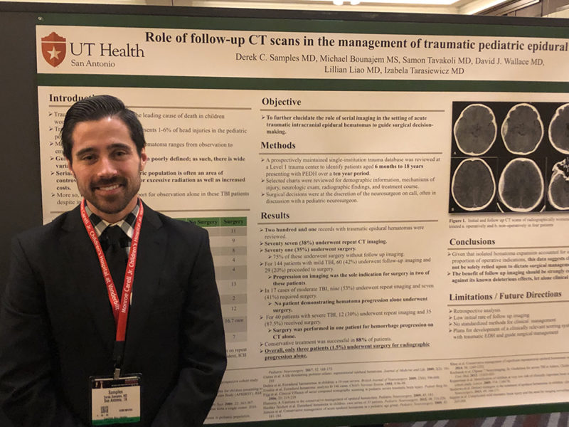 Dr. Derek Samples standing in front of his research poster and smiling.