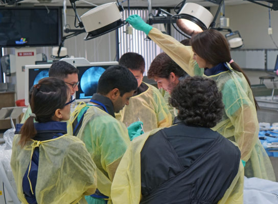 People in scrubs standing around the cadaver lab with Dr. Sun. One woman adjusts a giant lamp.