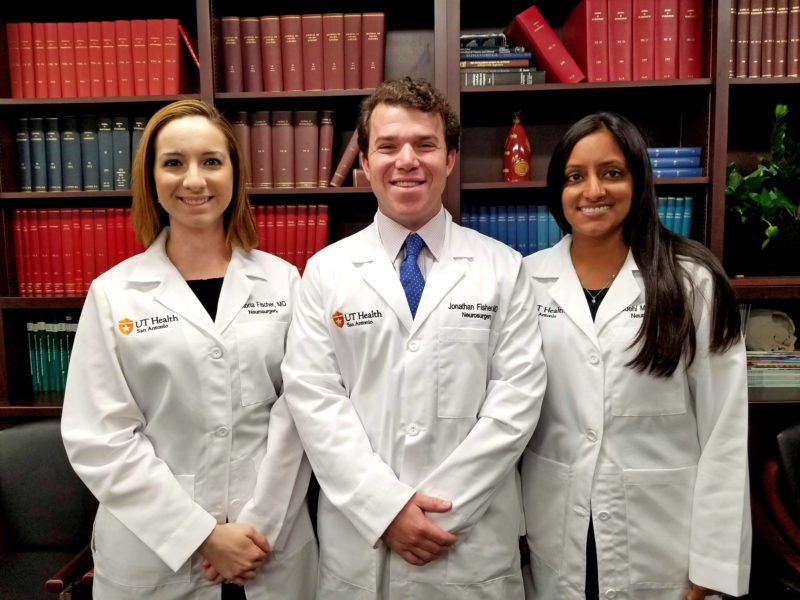 Drs. Victoria Fischer, Jonathan Fisher, and Vaidehi Mahadev smiling and wearing white coats in the Department of Neurosurgery conference room.