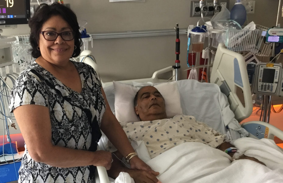 Patient Gustavo Macharro lying in a hospital bed while his wife stands by his side.