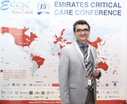 Dr. Ali Seifi smiling in front of a poster for the 15th Emirates Critical Care Conference.