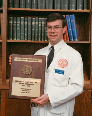 Dr. Fredrick Boop standing in a white coat with his graduation plaque.