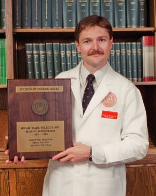 Dr. Micam Tullous standing in a white coat with his graduation plaque.