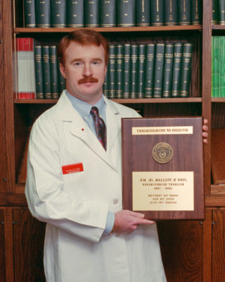Dr. John Pulliam standing in a white coat with his graduation plaque.