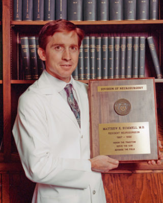 Dr. Matthew Hummell standing in a white coat with his graduation plaque.