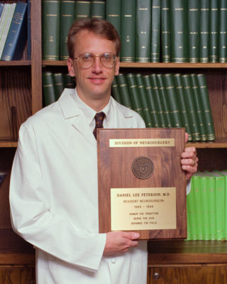 Dr. Daniel Peterson standing in a white coat holding his graduation plaque.