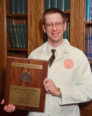 Dr. David Garrett standing in a white coat holding his graduation plaque.