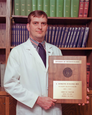 Dr. Dewayne Eubanks standing in a white coat holding his graduation plaque.