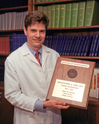 Dr. Christopher Gegg standing with a white coat holding his graduation plaque.