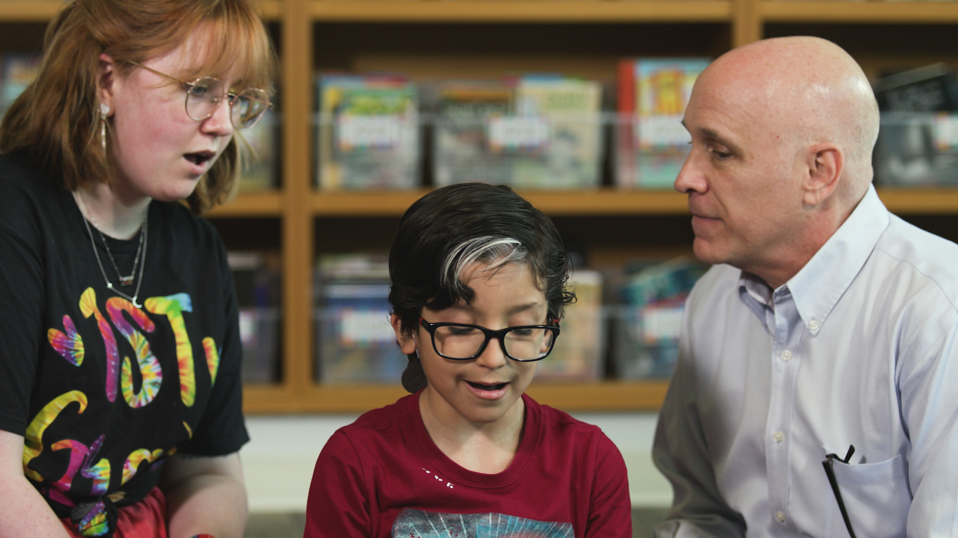 A deaf education student sits with a child who has a cochlear implant and the DEHS Program Director in front of an elementary school library stack