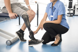 Male amputee with prosthesis in hospital. Man sitting on chair with nurse touching prosthetic limb. Female nurse working with male patient. Rehabilitation, recovery, improvement.